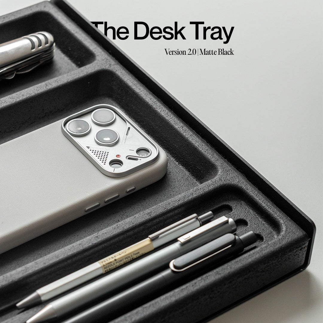 A black desk tray organizer shown on a light gray background, holding a smartphone, two pens, and a small tool. Text at the top reads 'The Desk Tray Version 2.