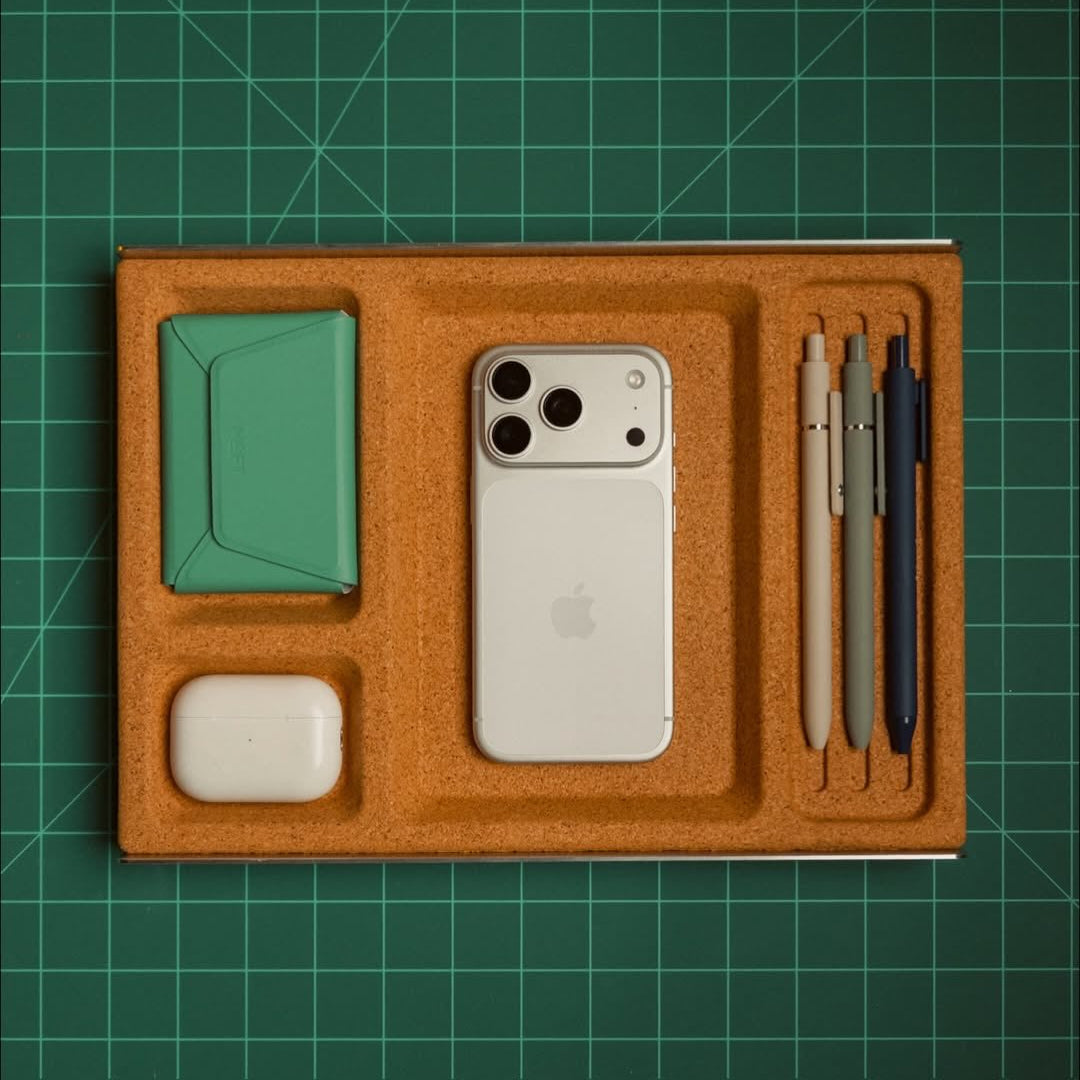 A cork organizer tray with a cutout layout, shown from above on a green cutting mat. The tray contains a white smartphone, a mint green card holder, a small white case, and three pens in beige, gray, and navy.